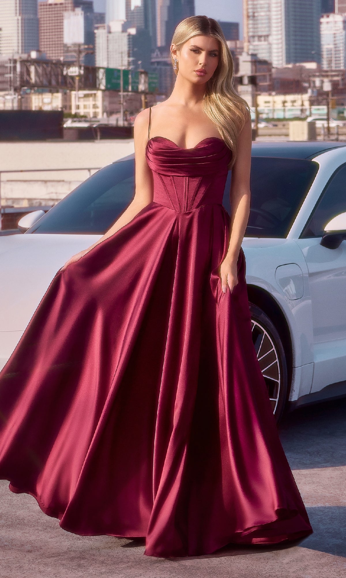 A woman wearing the Ladivine 7481 Long Satin A-Line Prom Dress stands outdoors in front of a white car, with a city skyline in the background.